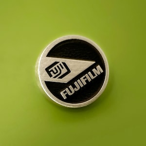 May include: A silver-colored Fujifilm logo emblem with a black center, set against a bright green background. The logo features the brand name and a stylized design.