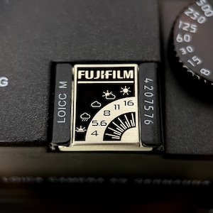 Op de afbeelding: Close-up van een zwarte camerabody met een Fujifilm-logo en belichtingsinstellingen. De instellingen omvatten cijfers en weersymbolen. De camera heeft ook een draaiknop met cijfers voor de sluitertijd.