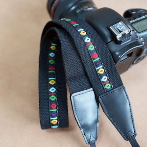 Könnte beinhalten: Schwarzer Kameragurt mit einem farbenfrohen geometrischen Muster in Rot, Grün, Gelb und Blau. Der Gurt ist an einer schwarzen DSLR-Kamera befestigt. Der Gurt hat schwarze Lederenden.