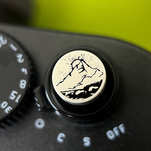 May include: Close-up of a black camera dial with white numbers and markings. A circular button features a mountain range design with a rising sun. The camera dial is set to "OFF".