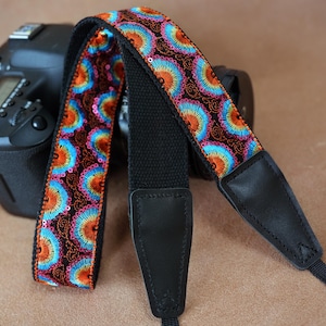 May include: A colorful camera strap with a floral pattern in orange, blue, and pink sequins on a black background. The strap is attached to a black camera body with a leather end piece.