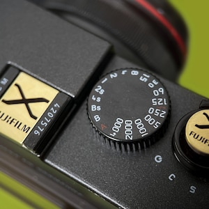 Puede incluir: Primer plano de una cámara Fujifilm negra con detalles dorados. La imagen destaca los diales, la configuración y el logotipo de Fujifilm. La cámara tiene una superficie texturizada y un fondo verde.