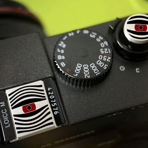 Puede incluir: Una parte superior de cámara negra con un botón disparador circular y una placa accesoria rectangular, ambos decorados con un patrón de líneas onduladas blancas y negras y un icono de ojo rojo central, junto a un dial de velocidad de obturación.