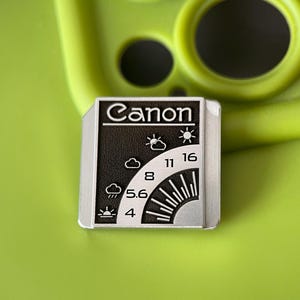 May include: A rectangular metal pin featuring a black and silver design with the word Canon at the top. The graphic displays a camera aperture scale with f-stop numbers 4, 5.6, 8, 11, and 16, alongside weather icons representing various lighting conditions.
