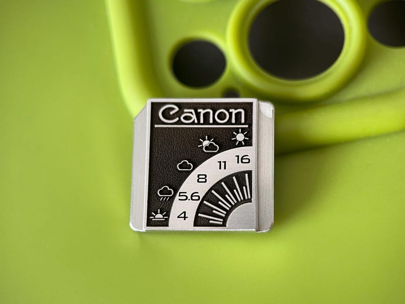 May include: A rectangular metal pin featuring a black and silver design with the word Canon at the top. The graphic displays a camera aperture scale with f-stop numbers 4, 5.6, 8, 11, and 16, alongside weather icons representing various lighting conditions.