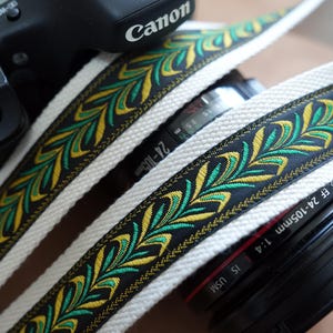 May include: Close-up of a camera with a decorative strap. The strap is white with a black border and a repeating pattern of green and yellow leaves. The camera lens has the text "EF 24-105mm 1:4 IS USM". The camera body has the Canon logo.