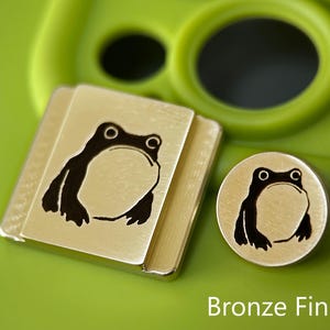 May include: Bronze frog-themed metal accessories. The image shows a square and a round piece, each with a black frog illustration. The text "Bronze Finish" is visible at the bottom. The background is a bright green.