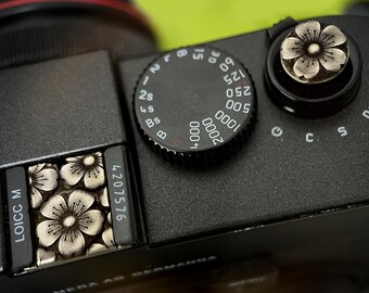 May include: Close-up of a black camera body with silver floral accents. The camera features a dial with numbers and settings, and a decorative flower design on the shutter button and a compartment. The serial number 4207576 is visible.