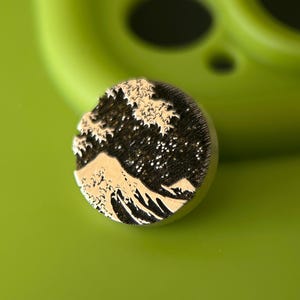Pode incluir: Um alfinete redondo de metal com uma representação detalhada de "A Grande Onda de Kanagawa" de Hokusai. O design mostra uma onda estilizada, montanha e céu em preto e branco sobre um fundo escuro. O alfinete está sobre uma superfície verde.