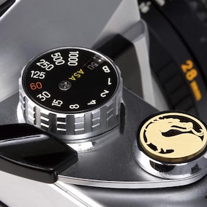 May include: A silver camera with a black dial that reads "ASA" and has numbers from 6 to 1000. The dial is set to 100. A black lever is visible on the left side of the camera. A gold-colored round button with a dragon design is on the right side of the camera.