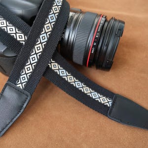 May include: Black camera strap with a geometric diamond pattern in white and light blue. The strap is attached to a black camera body and lens, resting on a brown surface. The strap has black leather ends.
