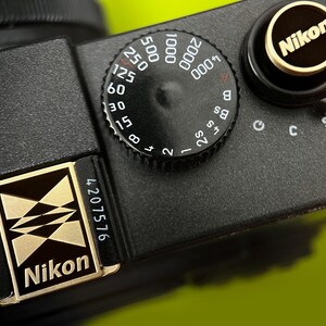May include: Close-up of a black Nikon camera with a gold and black logo. The camera features a dial with numbers and settings, including 250, 125, 60, 30, 15, 8, 4, 2, B, and 4000. The camera also has a serial number.