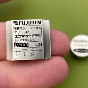May include: A silver metal film canister with the text "FUJIFILM", "業務用カラーフイルム", "アリントは", "ユニカカラー 2023/6/2", "をおすすめします.", "LV100", "135-24", "Fully", "Parlaty", "Unexposed", "Exposed", "Exposed", "Processed", "ODXI", "Noies" printed on it.