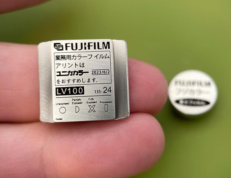 May include: A silver metal film canister with the text "FUJIFILM", "業務用カラーフイルム", "アリントは", "ユニカカラー 2023/6/2", "をおすすめします.", "LV100", "135-24", "Fully", "Parlaty", "Unexposed", "Exposed", "Exposed", "Processed", "ODXI", "Noies" printed on it.
