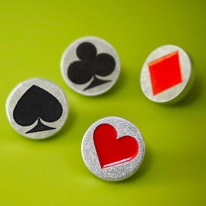 May include: Four round silver-toned buttons with playing card suit symbols: a black spade, a black club, a red diamond, and a red heart. The buttons are set against a bright green background.