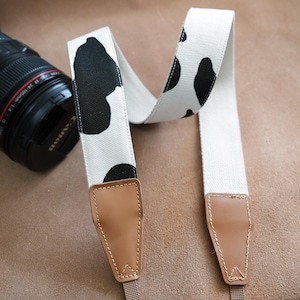 May include: A camera strap featuring a white fabric with black cow-like spots. The strap has brown leather ends and is next to a black Canon camera.