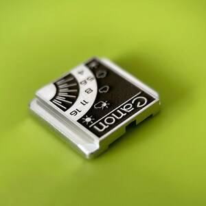 May include: A square silver-toned metal accessory featuring a black and white graphic panel. The panel displays the word Canon in white text, alongside a sun icon, cloud icons, and a numerical scale ranging from 4 to 16 with radial lines.