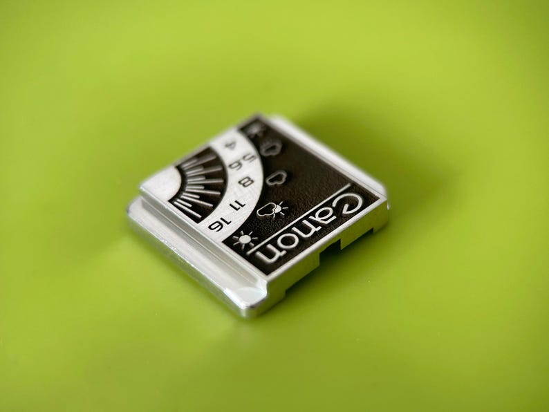 May include: A square silver-toned metal accessory featuring a black and white graphic panel. The panel displays the word Canon in white text, alongside a sun icon, cloud icons, and a numerical scale ranging from 4 to 16 with radial lines.