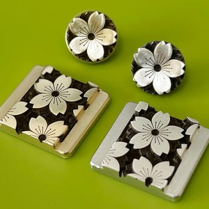 Op de afbeelding: Zilverkleurige metalen stempels en knopen met een kersenbloesem ontwerp. Twee vierkante stempels en twee ronde knopen zijn gerangschikt op een groene ondergrond. De bloesems hebben witte bloemblaadjes en een zwart centrum.