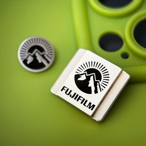 Könnte beinhalten: Zwei kleine, silberfarbene Fujifilm-Logo-Anstecknadeln mit Berg- und Sonnendesign. Eine ist rund, die andere quadratisch. Die Anstecknadeln liegen auf einer hellgrünen Oberfläche, in der Nähe einer grünen Handyhülle.