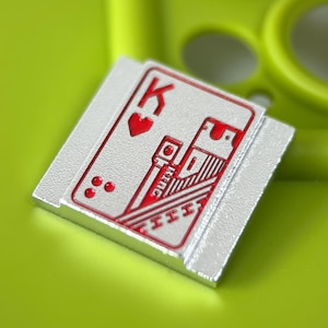 May include: A silver-colored, rectangular playing card with a red King of Hearts design. The card features the letter "K" and the word "King" in red, along with red heart and dot symbols. The card is set against a bright green background.