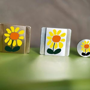 May include: Three metal objects with a sunflower design. The sunflowers have yellow petals, orange centers, and green leaves. The objects are a square, a rectangular square, and a small circle.