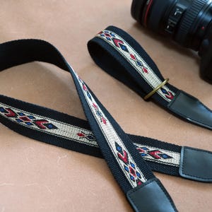 Könnte beinhalten: Schwarzer Kameragurt mit einem gewebten Muster in Rot, Blau und Creme. Der Gurt hat schwarze Lederenden und einen Metallring. Im Hintergrund ist eine schwarze Kamera zu sehen.
