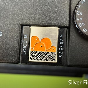 May include: Silver camera accessory with a rectangular design. The accessory has a silver background with an orange graphic of three round shapes. The bottom of the design has a black and white dotted pattern. The camera body is black.