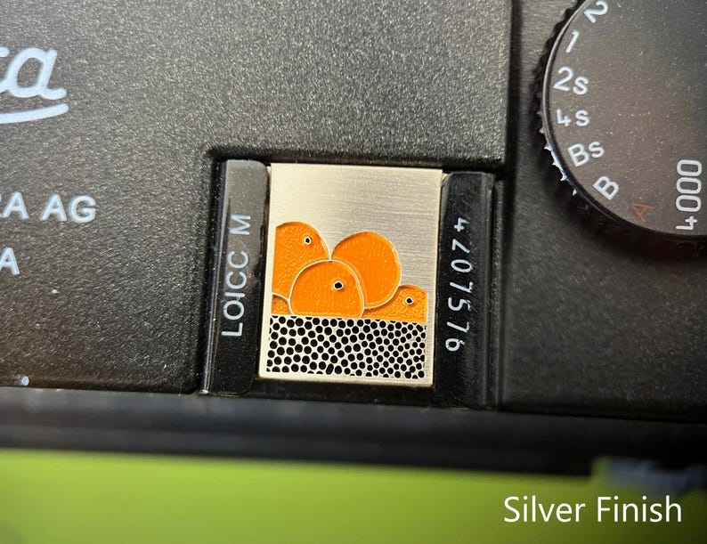 May include: Silver camera accessory with a rectangular design. The accessory has a silver background with an orange graphic of three round shapes. The bottom of the design has a black and white dotted pattern. The camera body is black.