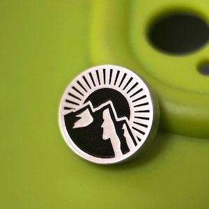 Könnte beinhalten: Ein runder, silberfarbener Metallpin mit einem schwarzen Berg- und Sonnendesign. Der Pin zeigt eine Bergsilhouette mit einem Sonnenstrahlenmuster. Der Pin befindet sich auf einem hellgrünen Hintergrund.