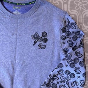 May include: A light blue sweatshirt with a black floral design on one sleeve. The sweatshirt has a green tag that says "Tek Gear" on the neck.
