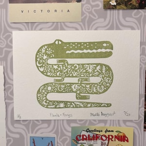 May include: A green print of an alligator with floral and fauna details. The artwork is titled "Flomis + Fangs" by Molli Suygast. The print is on white paper and is displayed with other postcards and ephemera.