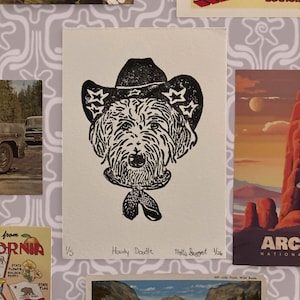 Peut inclure: Estampe en linogravure noir et blanc d'un chien portant un chapeau de cow-boy et un bandana. L'estampe est intitulée "Howdy Doodle" et signée par l'artiste. L'estampe est affichée avec d'autres cartes postales et œuvres d'art.