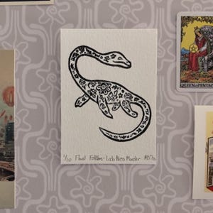 Può includere: Illustrazione in bianco e nero del mostro di Loch Ness con motivi floreali. L'opera d'arte è su una carta bianca con il testo "1/10 Floral Folklore-Lach Ness Monster MS5/8". La carta è esposta con altre cartoline.