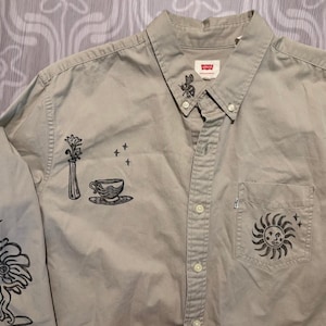 Op de afbeelding: Een licht olijfgroen button-down shirt met zwarte grafische illustraties. Het shirt heeft een zonontwerp op de zak, bloemen- en theekopjesontwerpen en een Levi's label. Het shirt met lange mouwen heeft een button-down kraag.