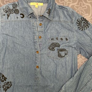 May include: A light blue denim button-down shirt featuring black ink-style illustrations of a croissant, a coffee mug, a sun, a crescent moon, and floral sprigs. The shirt has a collar, a front chest pocket, and brown buttons down the center.