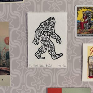 Può includere: Illustrazione in bianco e nero di Bigfoot con motivi floreali. Il testo "1/12 Floral Folklore-Bigfoot MS 1/25" è visibile. L'immagine è circondata da altre cartoline e oggetti effimeri.