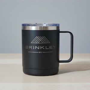 May include: Black travel mug with a silver rim and handle. The mug has a clear lid and features the text "BRINKLEY RV" with a mountain graphic. The mug is designed to keep beverages hot or cold.