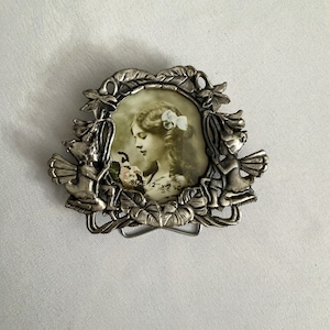 May include: Silver-toned metal belt buckle with a vintage-style oval picture of a woman with flowers. The buckle is decorated with leaves and two cherub figures. The picture is 2" x 2".