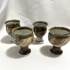 May include: Four ceramic cups with a brown and green glaze. Each cup has a unique leaf design and a short stem.