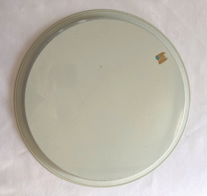 May include: A white metal serving tray with a slightly raised rim. The tray has a smooth surface and a small, blue and brown sticker on the side.