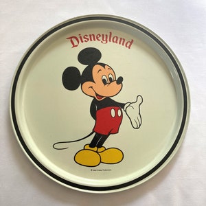 May include: A white metal serving tray with a black rim. The tray features a cartoon image of Mickey Mouse in red shorts and yellow shoes. The text "Disneyland" is printed in red letters above Mickey Mouse. The copyright text "&copy; Walt Disney Productions" is printed in black at the bottom of the tray.