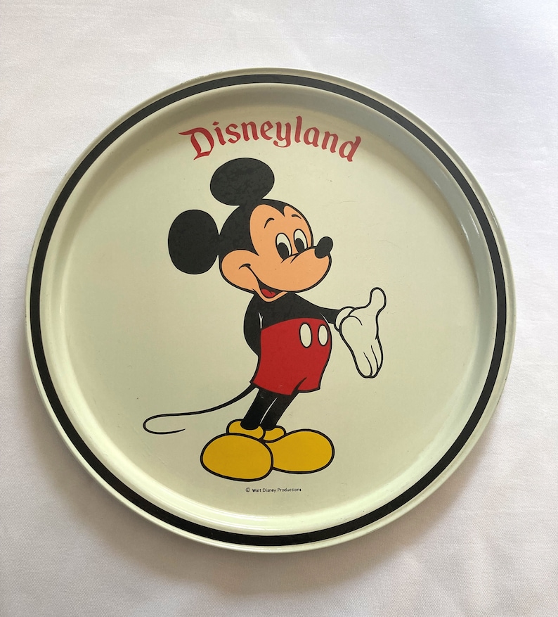 May include: A white metal serving tray with a black rim. The tray features a cartoon image of Mickey Mouse in red shorts and yellow shoes. The text "Disneyland" is printed in red letters above Mickey Mouse. The copyright text "&copy; Walt Disney Productions" is printed in black at the bottom of the tray.
