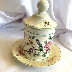 May include: A vintage ceramic sugar bowl with a lid and a matching saucer. The bowl and saucer are decorated with a floral pattern featuring pink, yellow, and green flowers and a green bird.