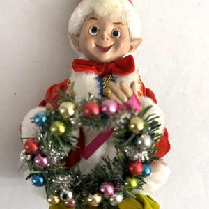 May include: A vintage Christmas elf doll with a smiling face, wearing a red coat with white trim, a red bow tie, and a yellow hat with pink trim. The elf holds a small wreath decorated with colorful ornaments.