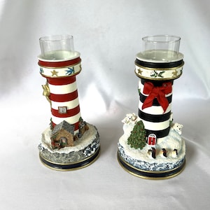 May include: Two white and red striped lighthouse candle holders with a winter scene. One has a red bow and the other has a small house. Both have a glass votive holder on top.