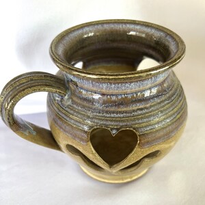 May include: A brown ceramic mug with a heart-shaped cutout and a light blue glaze. The mug has a handle and is perfect for holding a warm beverage.