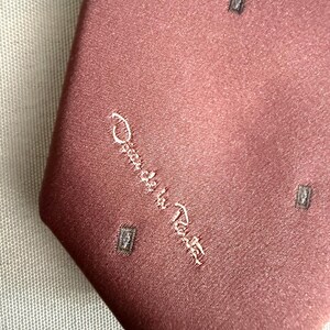 May include: A dusty rose-colored tie with a subtle sheen. The tie features a repeating pattern of small, dark gray squares and the embroidered signature "Oscar de la Renta" in a cursive script. The tie is made of a smooth fabric.