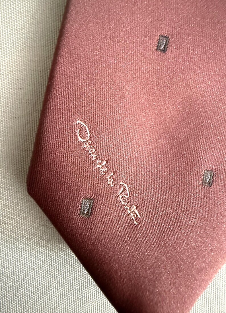 May include: A dusty rose-colored tie with a subtle sheen. The tie features a repeating pattern of small, dark gray squares and the embroidered signature "Oscar de la Renta" in a cursive script. The tie is made of a smooth fabric.