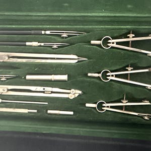 May include: An open, vintage drafting set in a green velvet-lined case. The set includes several silver-toned compasses, dividers, and other drawing tools. The case is black with a hinged lid, and the tools are neatly arranged.